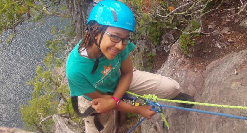 a person who is rock climbing above a body of water smiles at the camera